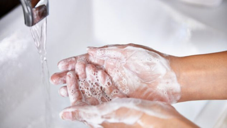 Woman washing her hands at the kitchen sink. There are vegetables out of focus in the background.